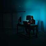 Hacker dressed in black and wearing a Guy Fawkes mask sits at a desk in a dark room, illuminated only up by the dim light of his laptop screen.